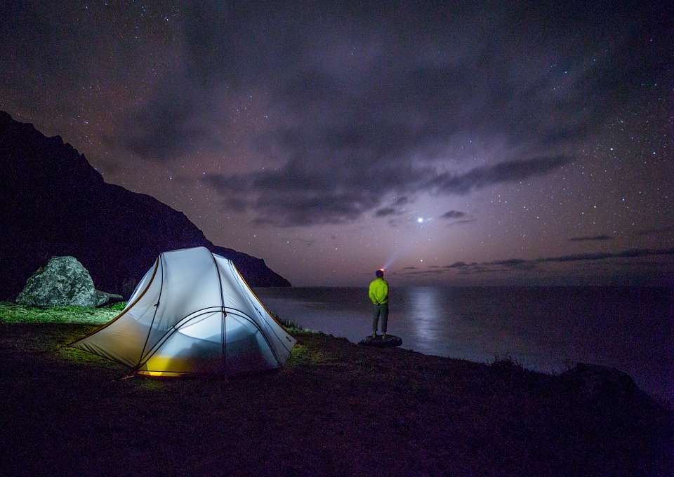 a tent with a light inside, a hiker standing with a headlight and looking into the night sky