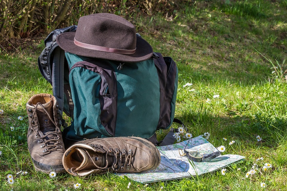 a backpack, a hat, a hiking shoes, a map and a compass on the ground with grasses and flowers