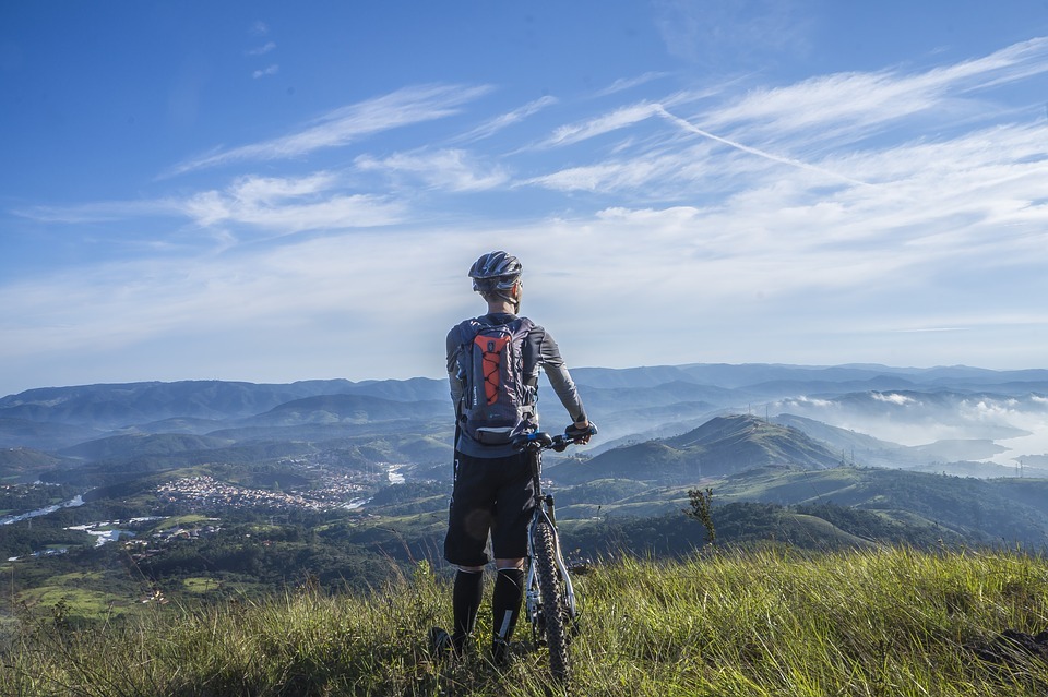 back of a person holding a bike, surface with green grass, blue sky partially covered with clouds, green hills