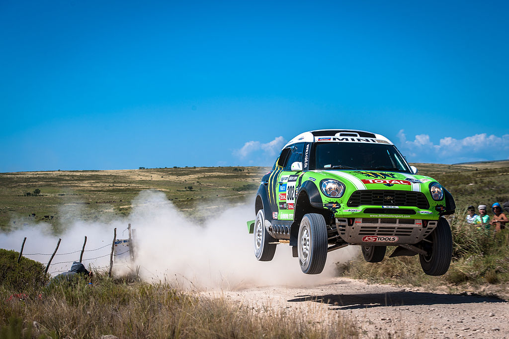 a green, off-road vehicle in mid-air, fence on the left, with dust cloud and a grassy field in the background
