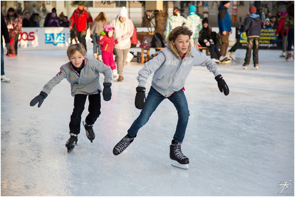 two boys ice skating, with people at their background wearing winter clothes and skating shoes
