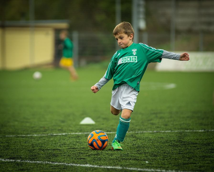 two children in a green jersey, child kicking the ball, green football field, wired fence