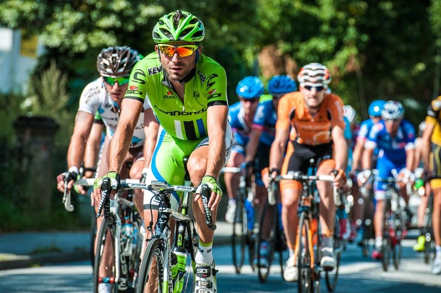 a cyclist in green jersey riding his bike, in the background is a blurred image of other cyclists, one in white, one in orange, and many others in blue jerseys